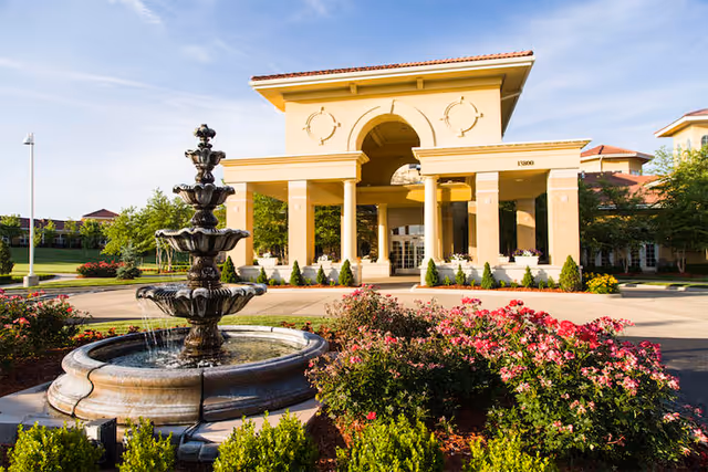 Exterior view of a senior living facility entrance with a fountain and landscaped gardens.