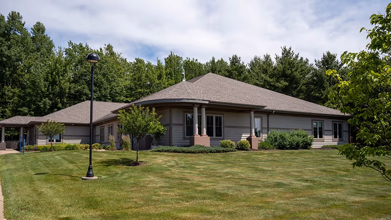 Single-story senior living facility exterior with a well-kept lawn, trees, and a lamppost at the front.
