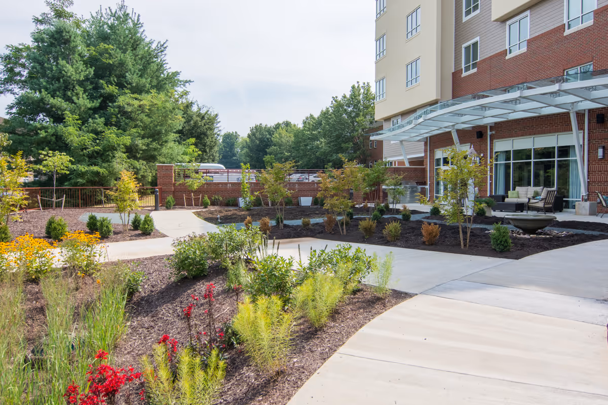 Landscaped outdoor courtyard and entrance of a multi-story assisted living building with walkways, plants, and seating under a canopy.