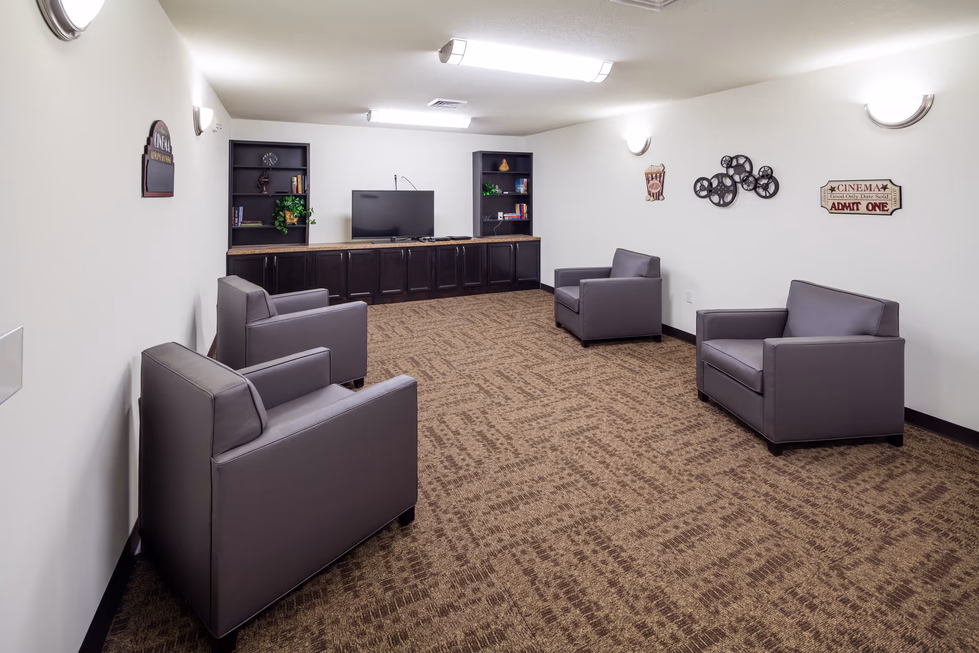 A small lounge room with four gray armchairs arranged around the perimeter. The room has beige patterned carpet, white walls with movie-themed decorations, and a TV on a dark wooden cabinet with shelves containing books and plants.