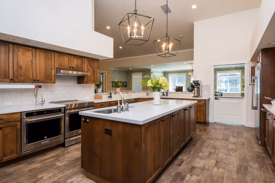 Bright modern kitchen with a large central island, wooden cabinets, stainless steel appliances, pendant lights, and a vase of flowers on the counter.