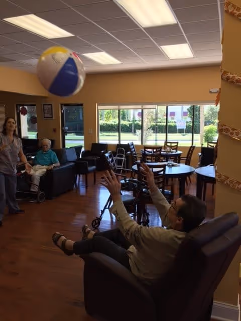 A group of seniors and a caregiver are playing with a beach ball in a bright common area with large windows, wooden floors, and several tables and chairs. One senior is seated in a recliner reaching up to catch the ball, while another senior sits on a couch and the caregiver stands nearby.