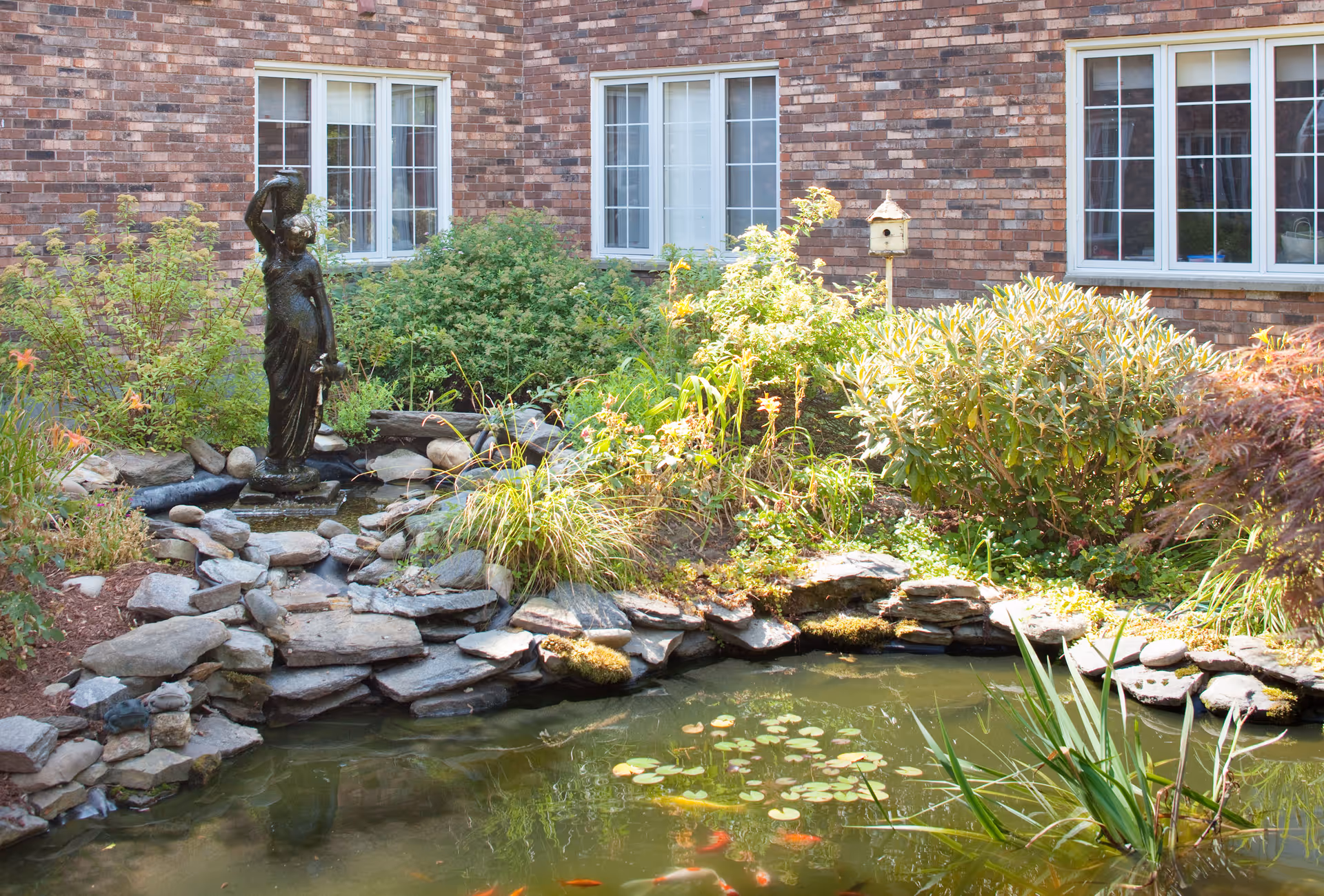 A tranquil outdoor garden area featuring a small pond with koi fish, surrounded by rocks and lush greenery. A bronze statue of a woman holding a jug stands near the pond, with a brick building and windows in the background.
