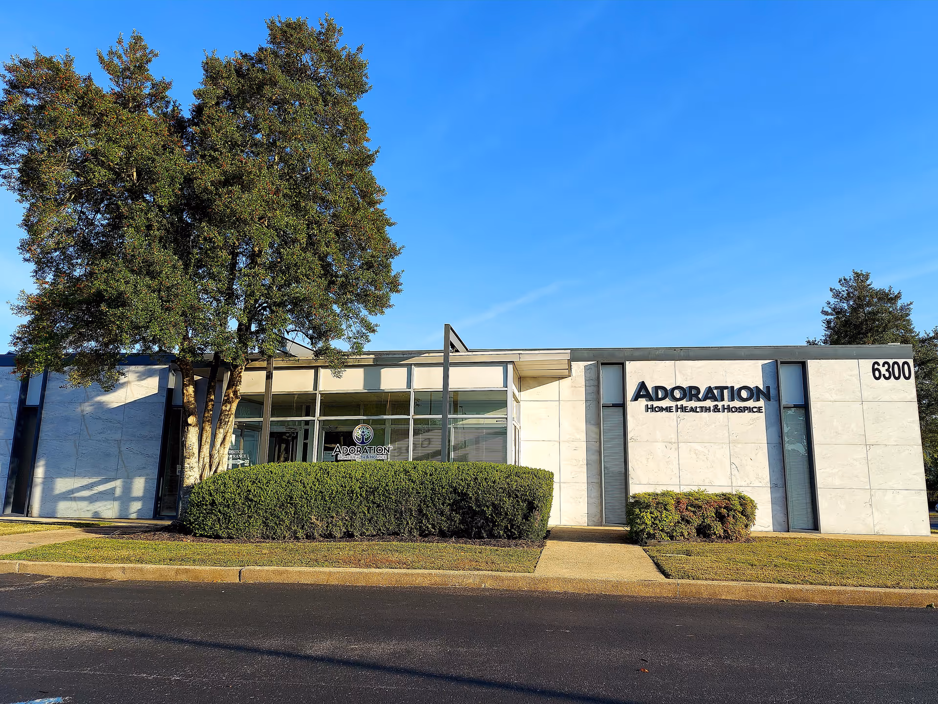 Modern single-story medical facility with Adoration Home Health and Hospice sign, trees, shrubs.