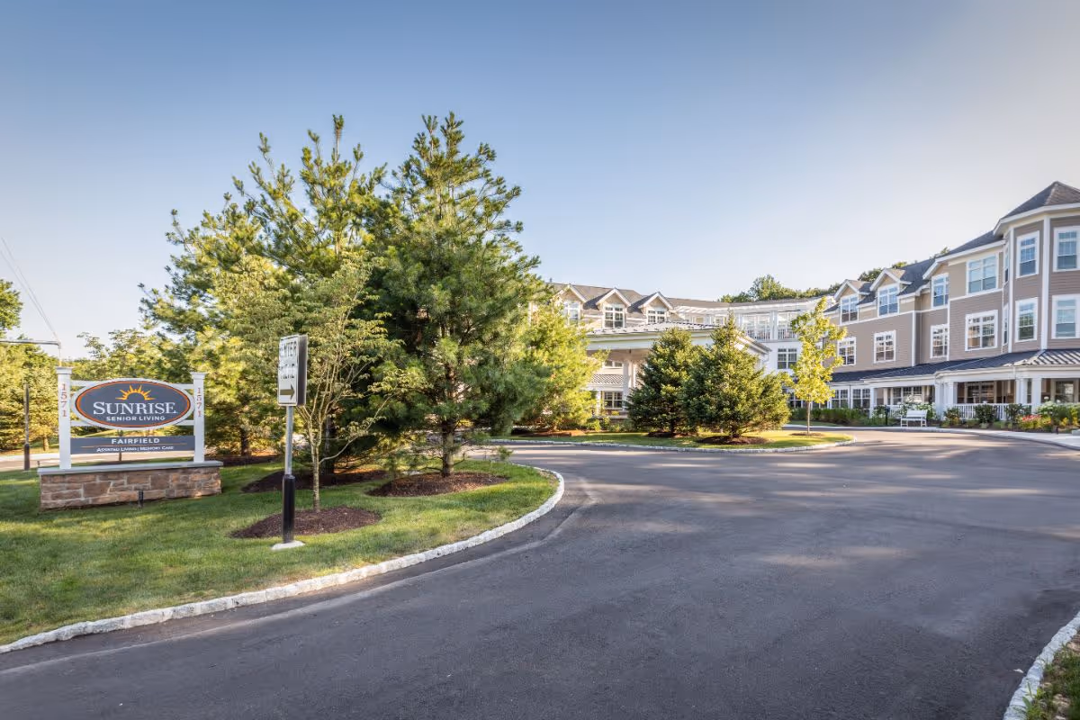 Exterior view of Sunrise of Fairfield senior living facility with a curved driveway, green trees, and a clear blue sky. The building is multi-story with many windows and a covered entrance. A sign with the facility name is visible on the left side near the road.
