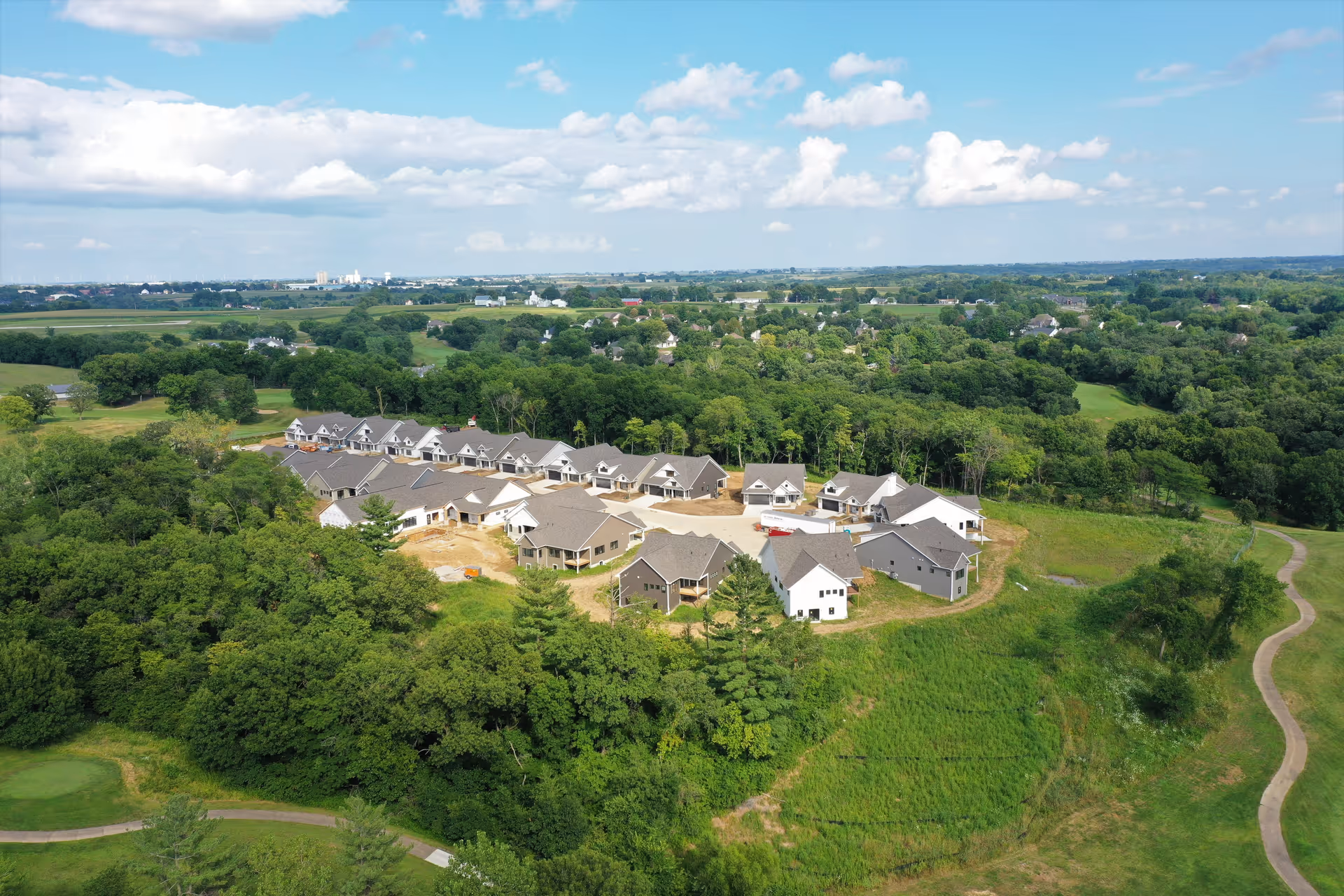 Aerial view of a small neighborhood of single-story residential buildings surrounded by trees and green fields.