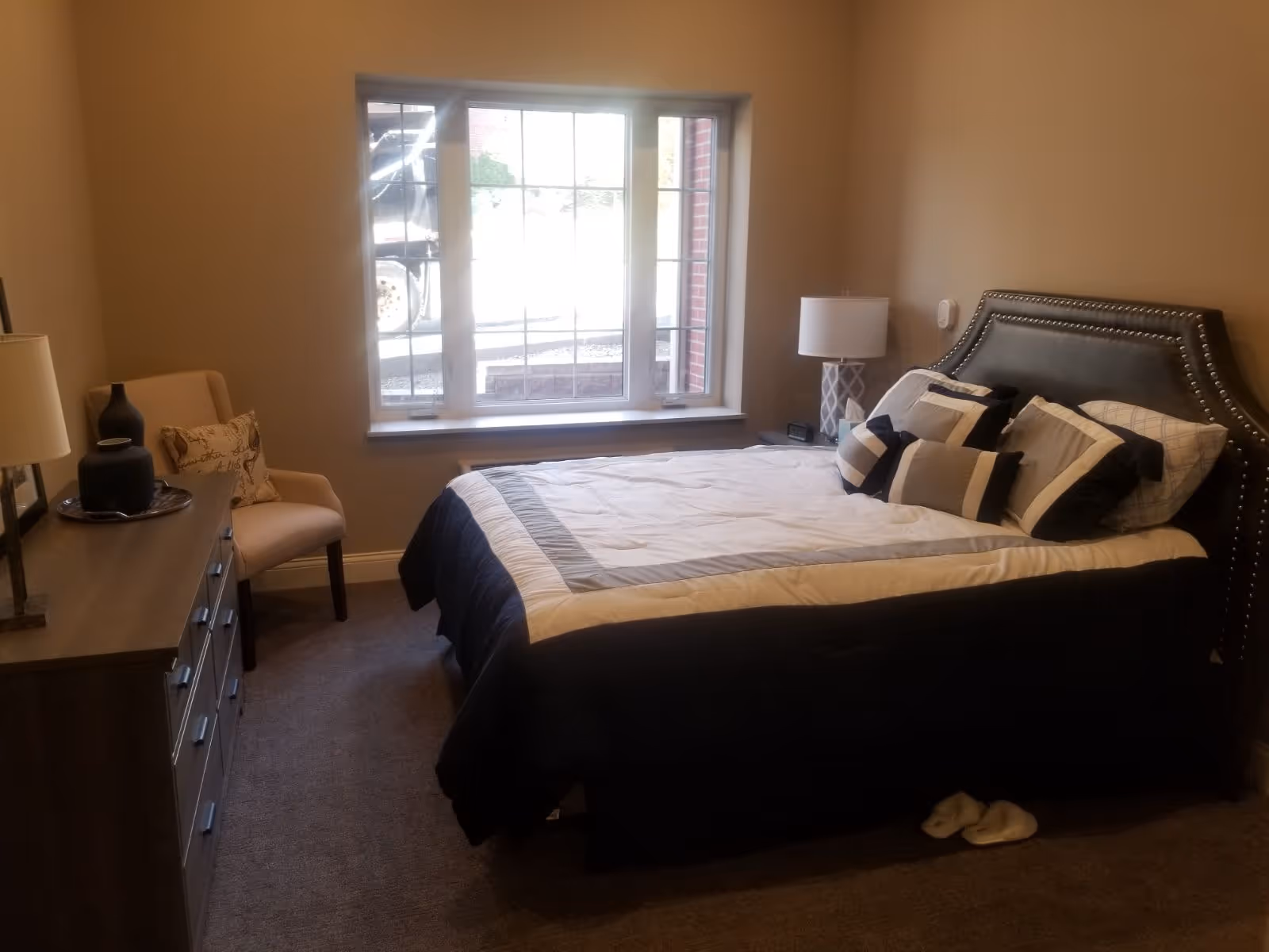 Neatly made bedroom with an upholstered headboard, bedside lamps, dresser, chair, and a large window.