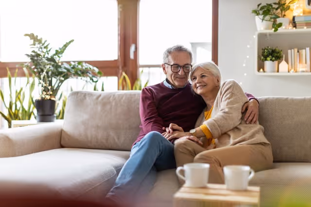 An elderly couple sitting closely together on a beige sofa in a cozy living room, smiling and holding hands. The room has large windows with natural light, potted plants, and shelves with books and decorative items.