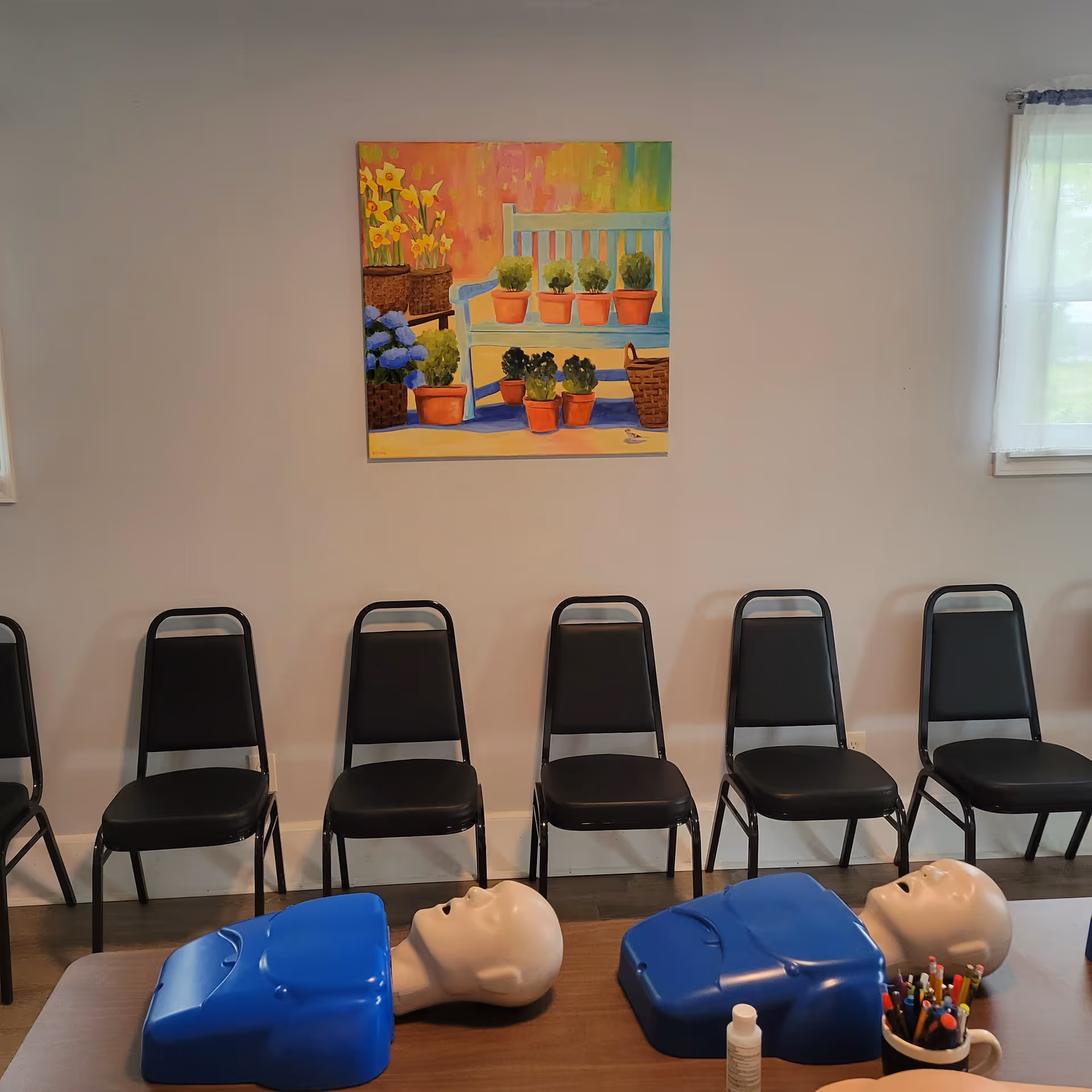 Row of black chairs against a wall with CPR training mannequins on a table.