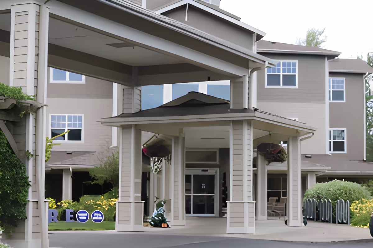 Entrance of a senior living facility named Cascades of Grants Pass with a covered driveway, beige exterior walls, multiple windows, and greenery around the building.