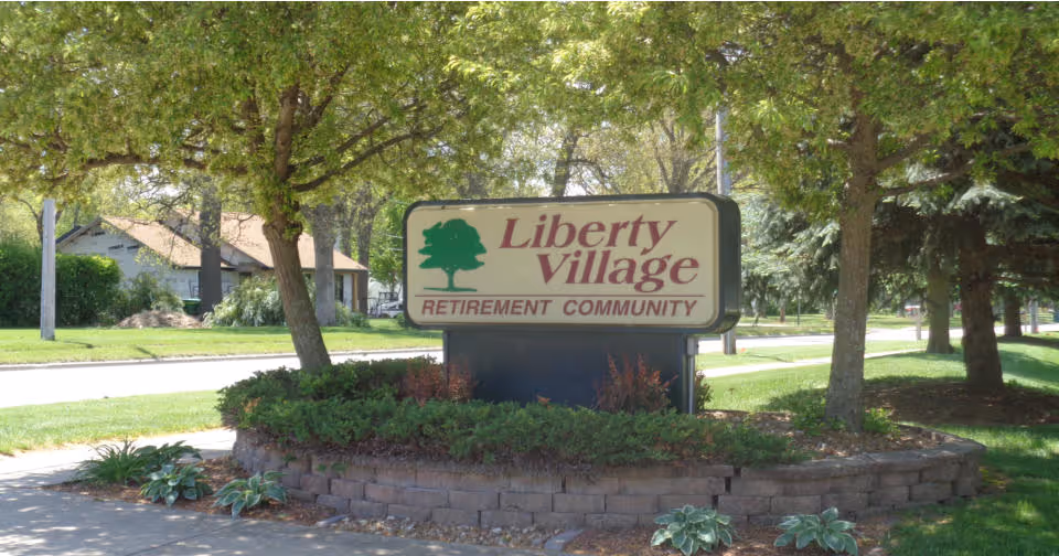 A landscaped roadside sign that reads "Liberty Village Retirement Community" beneath trees in front of residences.