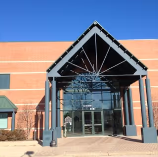 Brick senior living facility entrance with glass doors under a peaked metal canopy.