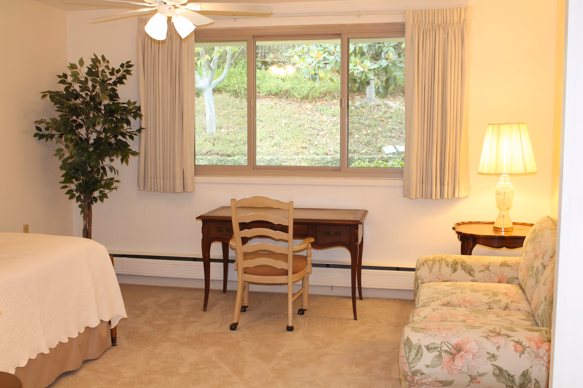 A cozy senior living bedroom with a bed covered in a white quilt, a wooden desk with a chair in front of a large window showing greenery outside, a floral patterned sofa, a side table with a lit lamp, and a tall potted plant in the corner.