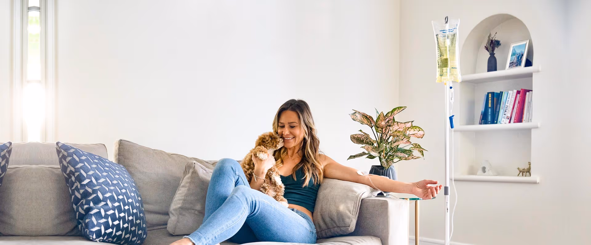 A woman sits on a gray sofa with a small dog in a bright living room.