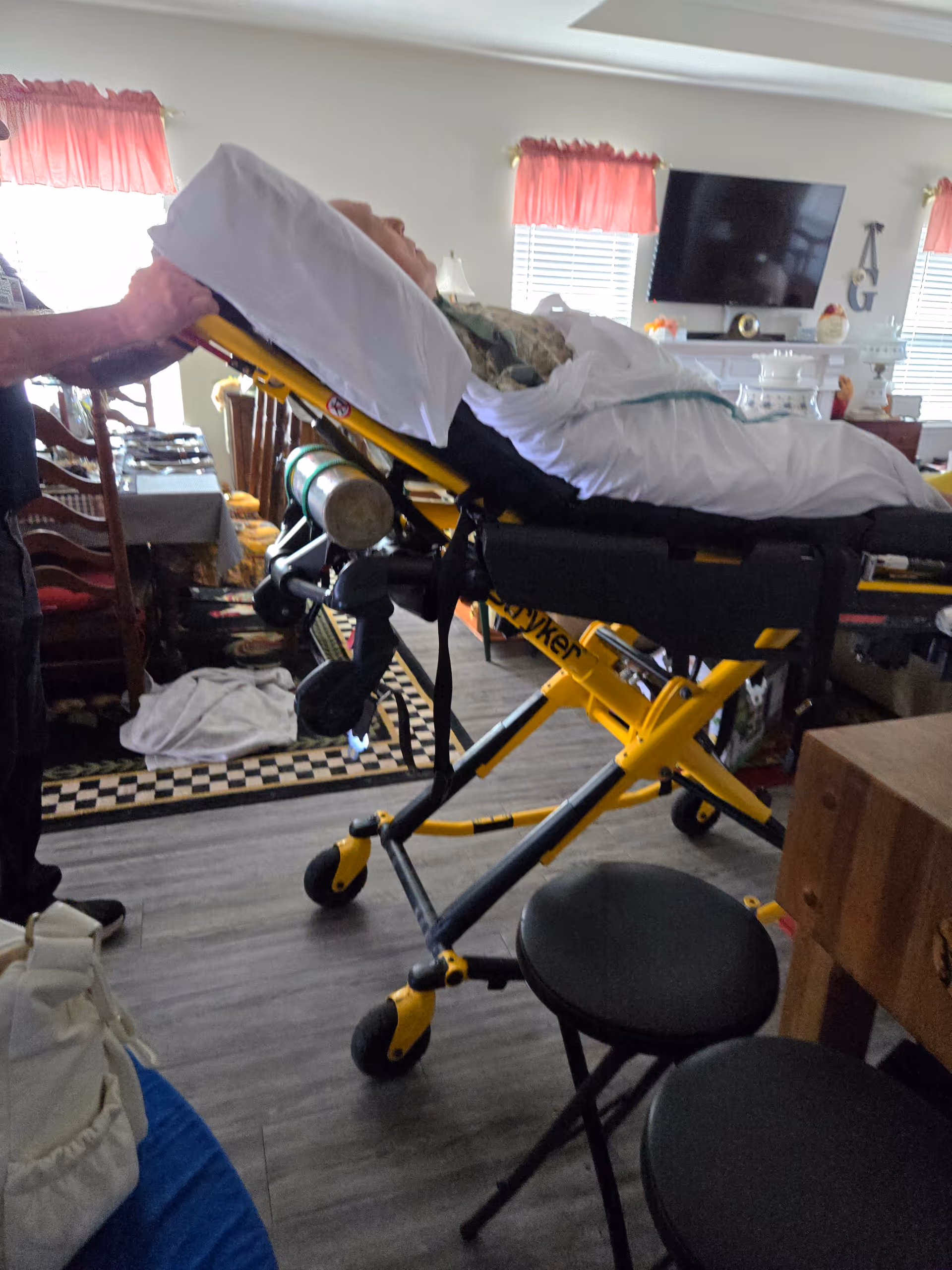 A medical stretcher stands in a furnished living room with chairs and a TV.