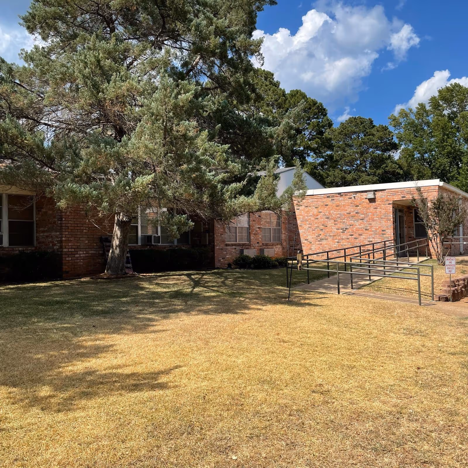 Exterior view of a brick building with a wheelchair accessible ramp leading to an entrance. There is a large tree casting shadows on the dry grass lawn in front of the building. The sky is partly cloudy with blue patches visible.