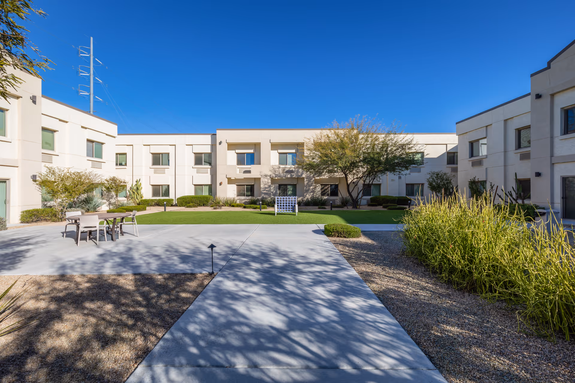 Outdoor courtyard area of BridgeWater Assisted Living - Deer Valley featuring a concrete walkway, green lawn, desert landscaping with bushes and trees, and a two-story beige building surrounding the courtyard under a clear blue sky.