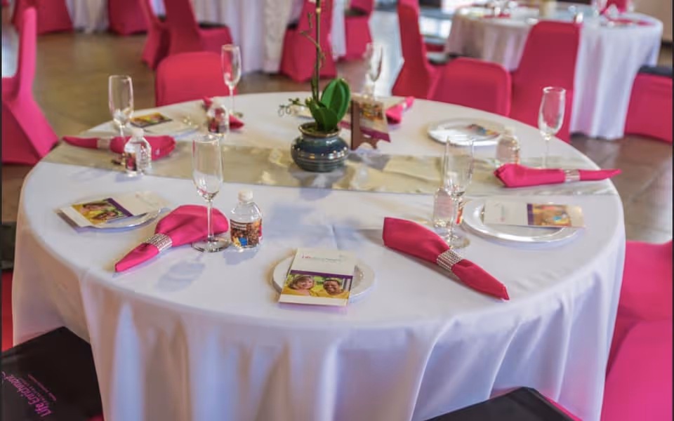 Round table with white cloth, pink napkins, glassware, and a central plant in a banquet setting.