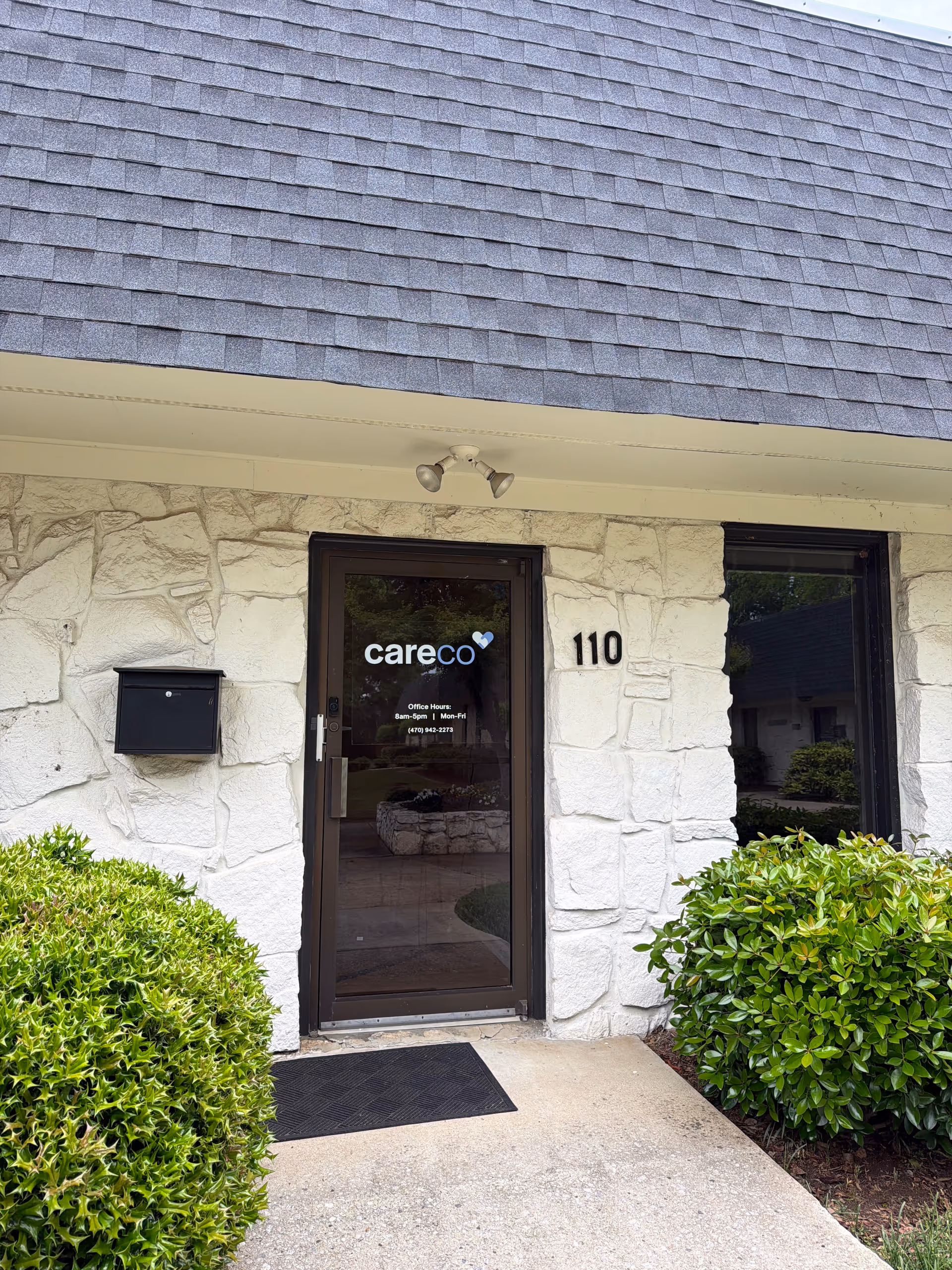 Stone exterior entrance with glass door labeled CareCo, number 110, surrounded by green shrubs.