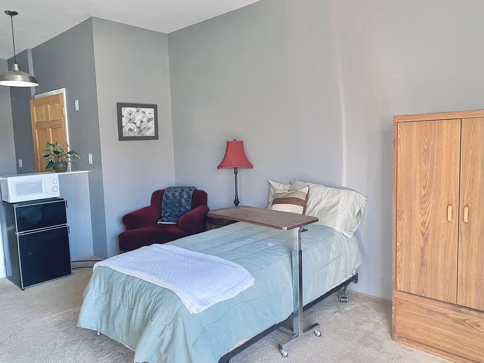 A bedroom in a senior facility with a bed, wardrobe, red chair, and a small kitchenette.