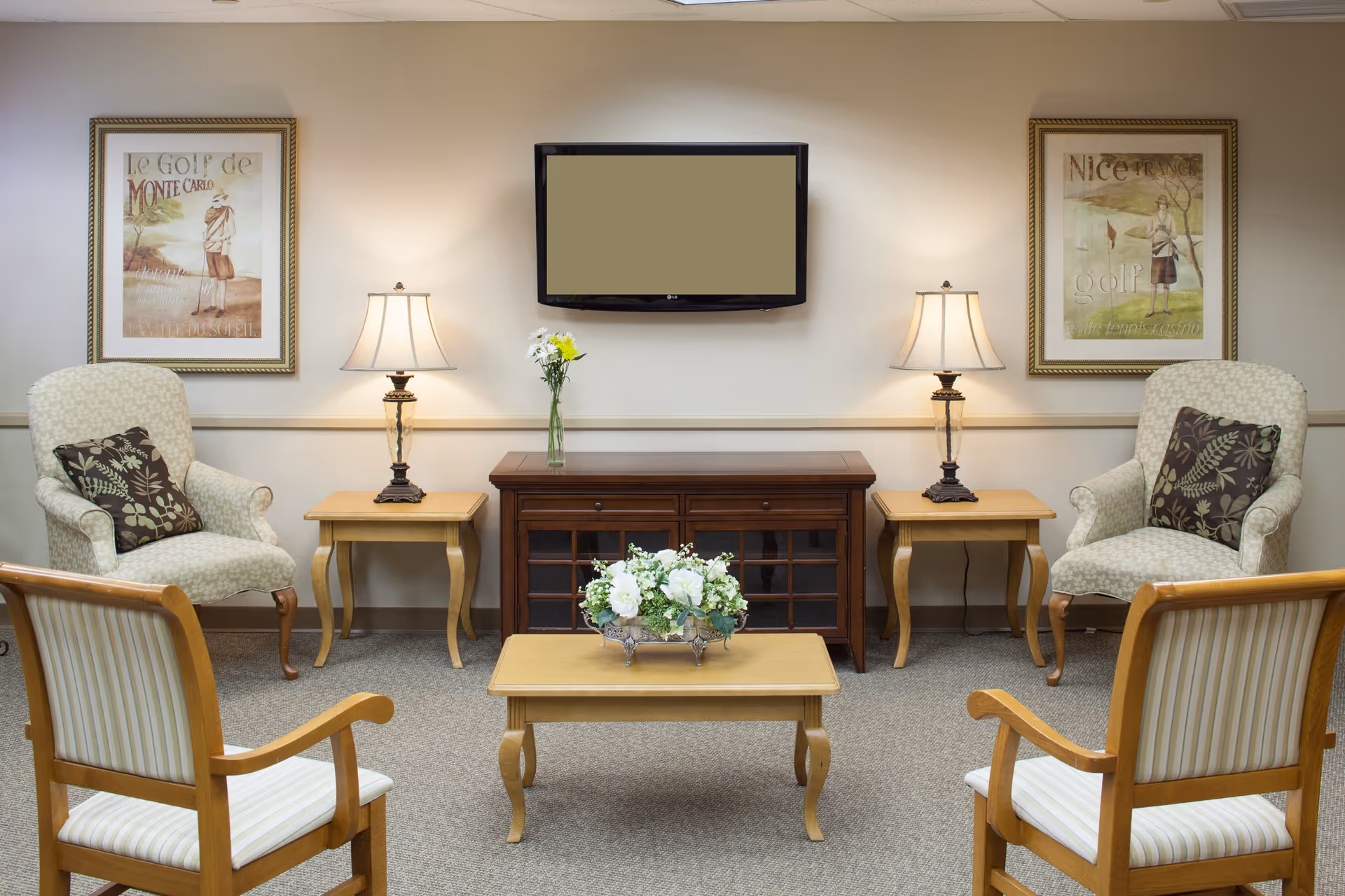 A cozy living room area with two upholstered armchairs with floral cushions, two wooden side tables each with a lamp, a wooden cabinet with a vase of flowers on top, a wall-mounted flat screen TV, two framed golf-themed pictures on the wall, and two wooden chairs with striped cushions facing the center coffee table with a floral arrangement.