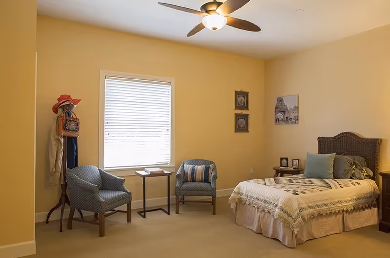 A softly lit bedroom with a bed, two armchairs by a window, a small side table, and a ceiling fan.
