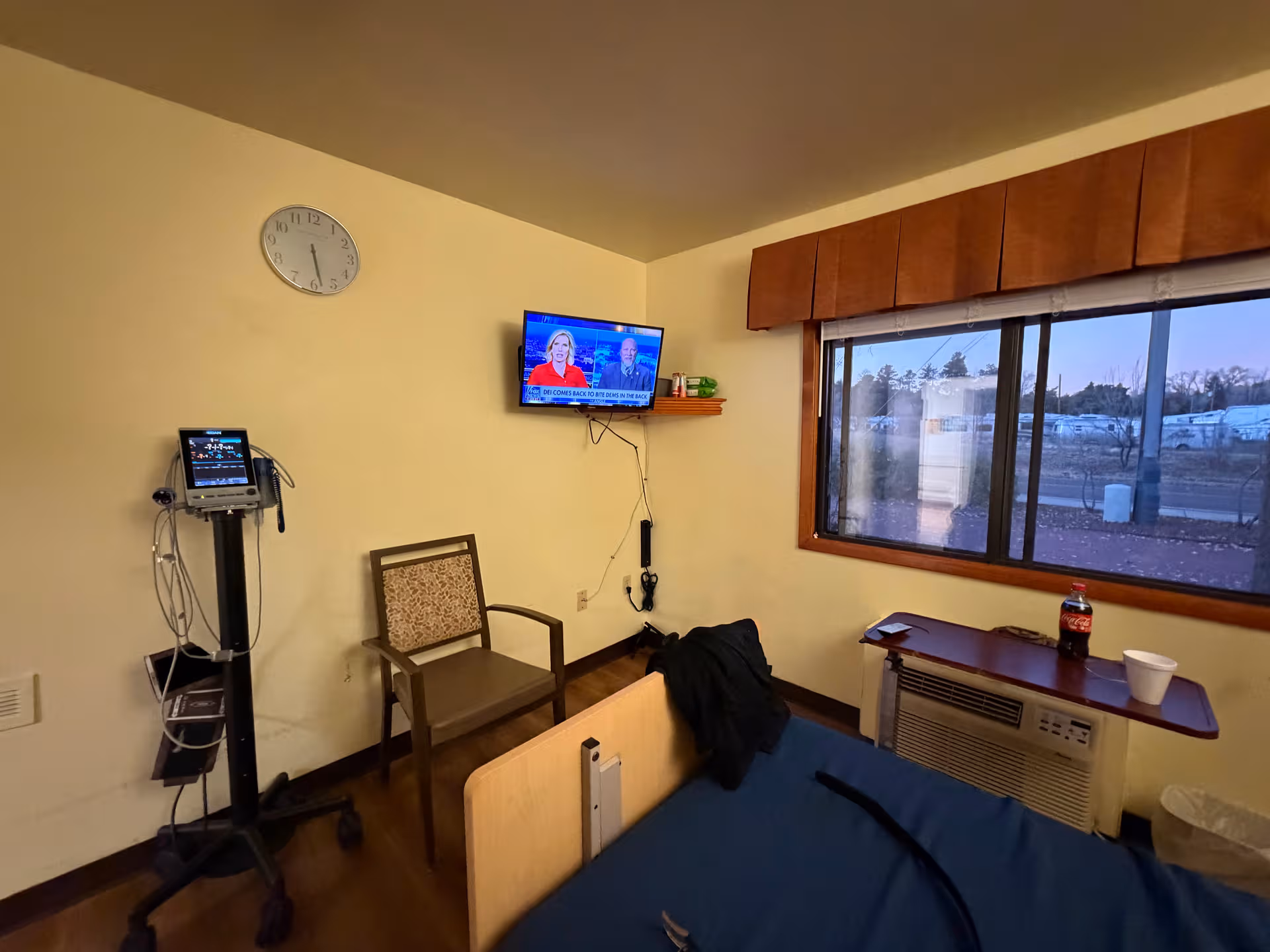A hospital-style patient room with a bed, medical monitor, chair, wall-mounted TV, window, and beverage table.
