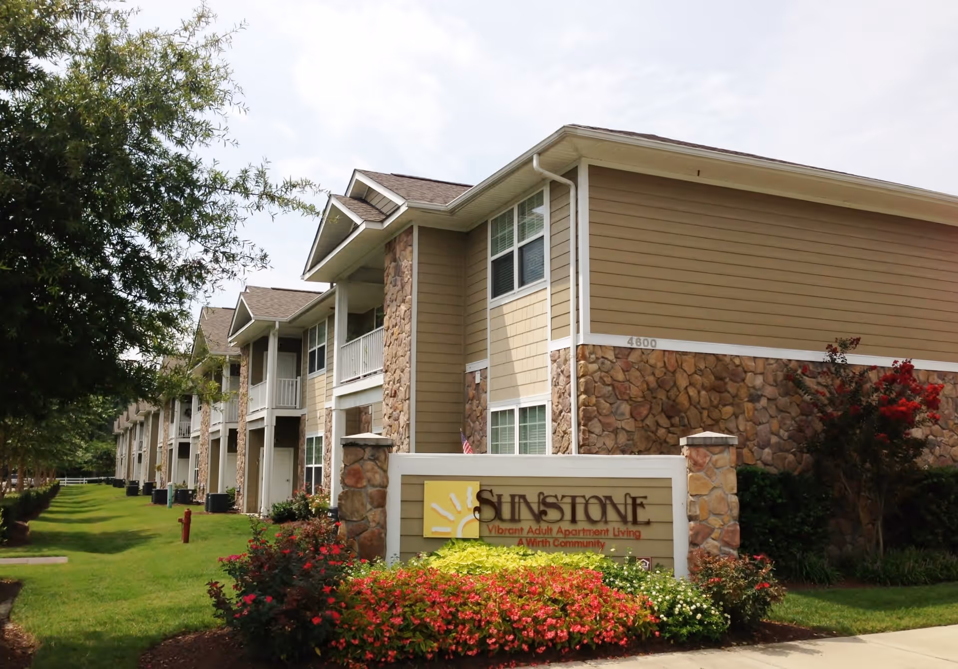 Exterior view of Sunstone Apartments, a two-story building with beige siding and stone accents. The building is surrounded by green grass, trees, and colorful flower beds. A sign in front reads 'Sunstone Vibrant Adult Apartment Living A Wirth Community.'