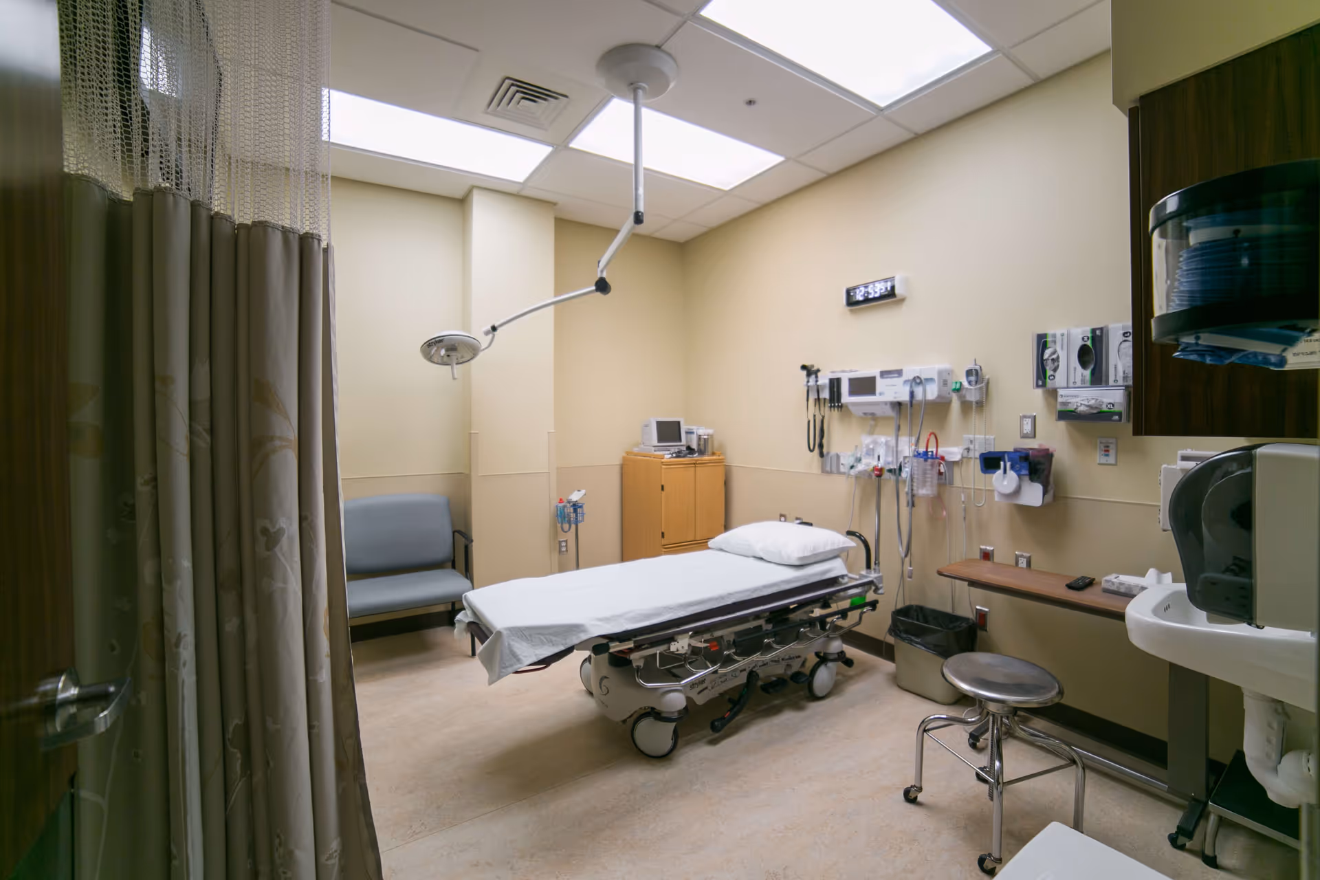 A medical examination room with a hospital bed covered with a white sheet and pillow, a gray chair against the wall, medical equipment mounted on the wall, a small wooden cabinet, a stool on wheels, a sink, and a privacy curtain partially drawn.