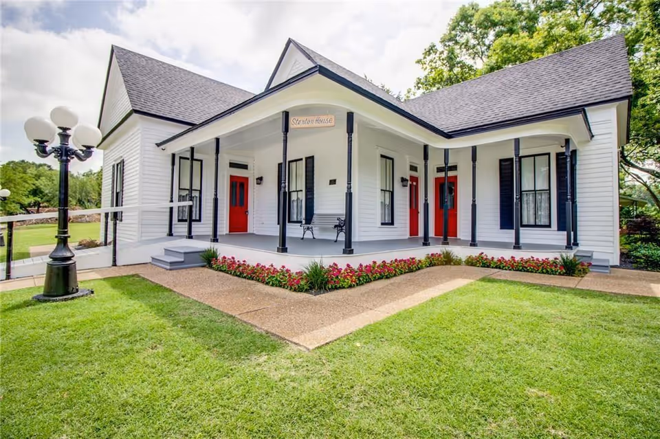 White single-story building with a wraparound porch, black columns, red doors, and landscaped lawn.