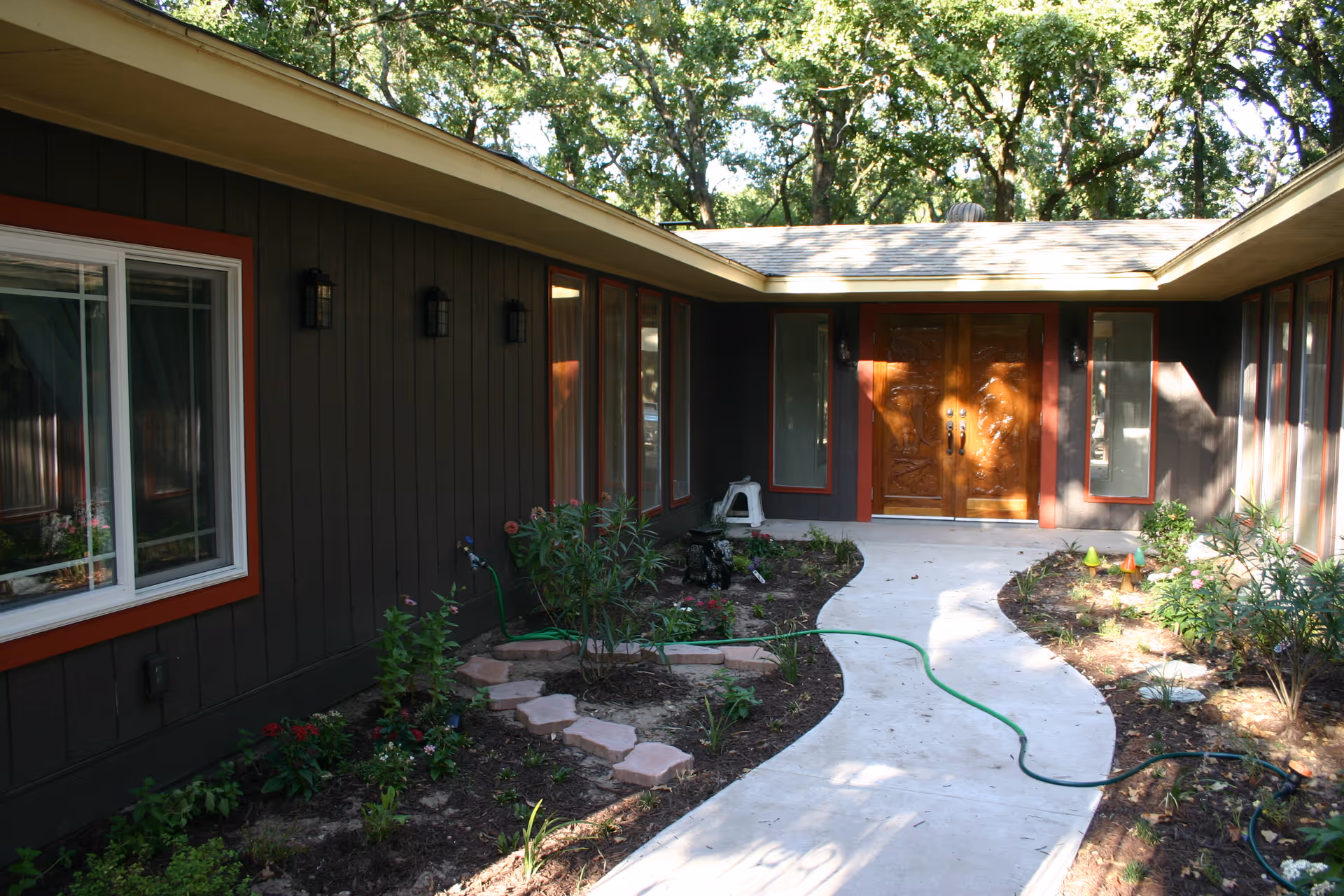 Exterior view of a single-story facility with dark siding, red trim, curved walkway, and garden beds.