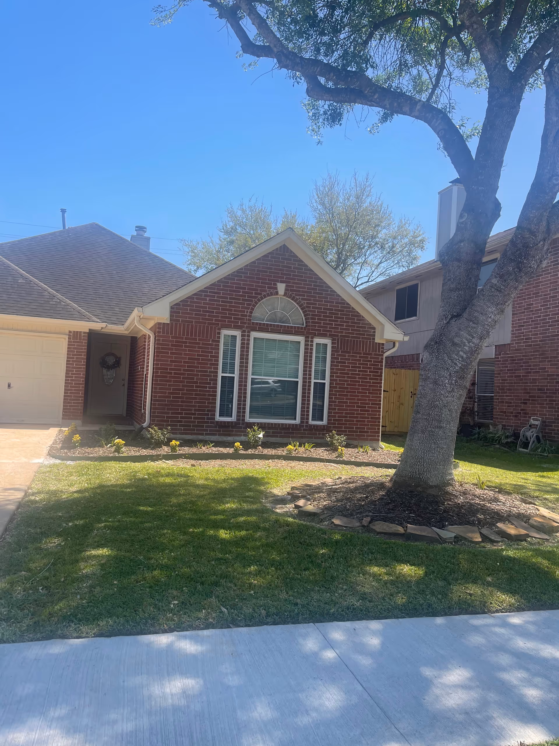 Brick single-story house front with an arched window, lawn, large tree, and driveway under a clear blue sky.