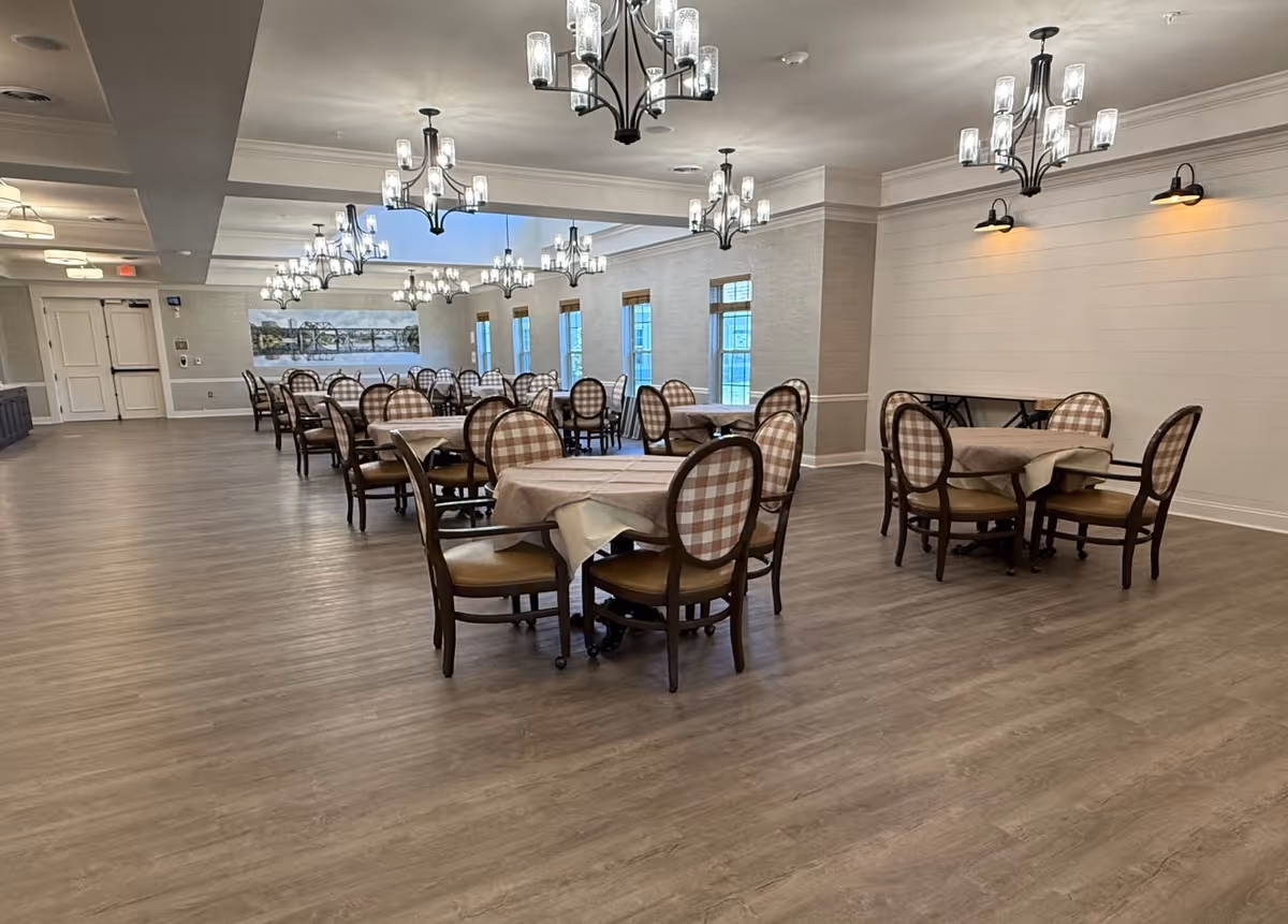 A spacious dining room with multiple round tables covered with beige tablecloths. Each table is surrounded by four wooden chairs with cushioned seats and backs featuring a checkered pattern. The room has wooden flooring, several large windows along one wall, and multiple modern chandeliers hanging from the ceiling providing ample lighting. The walls are light-colored with some wall-mounted lights and a large landscape painting is visible in the background.
