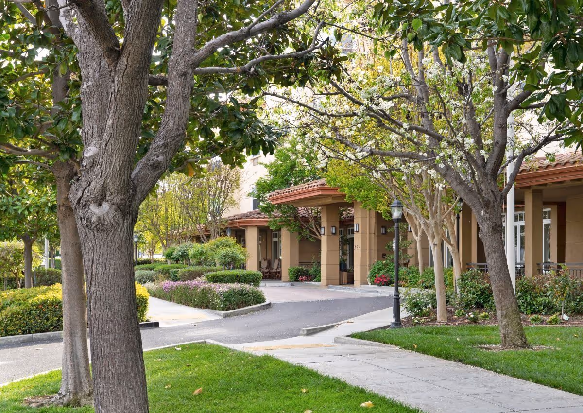 Landscaped front entrance with trees, shrubs, a sidewalk, and a covered drive-up area.