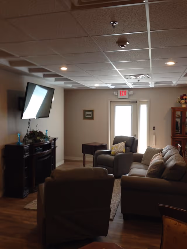 A cozy living room area with a mounted flat-screen TV above a dark wooden fireplace mantel. The room has a beige couch with multiple pillows, a matching armchair with a yellow patterned pillow, a small wooden side table, and a glass-front cabinet with flowers on top. There is an exit door with a window letting in natural light, and the ceiling has recessed lighting and a drop ceiling design.