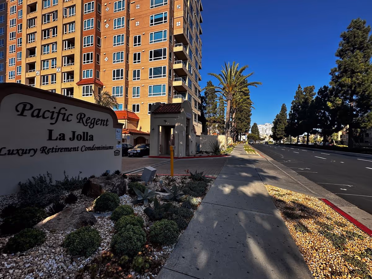 Street-level view of the Pacific Regent La Jolla retirement condominium building and entrance with sign, sidewalk, palm trees, and a nearby road.