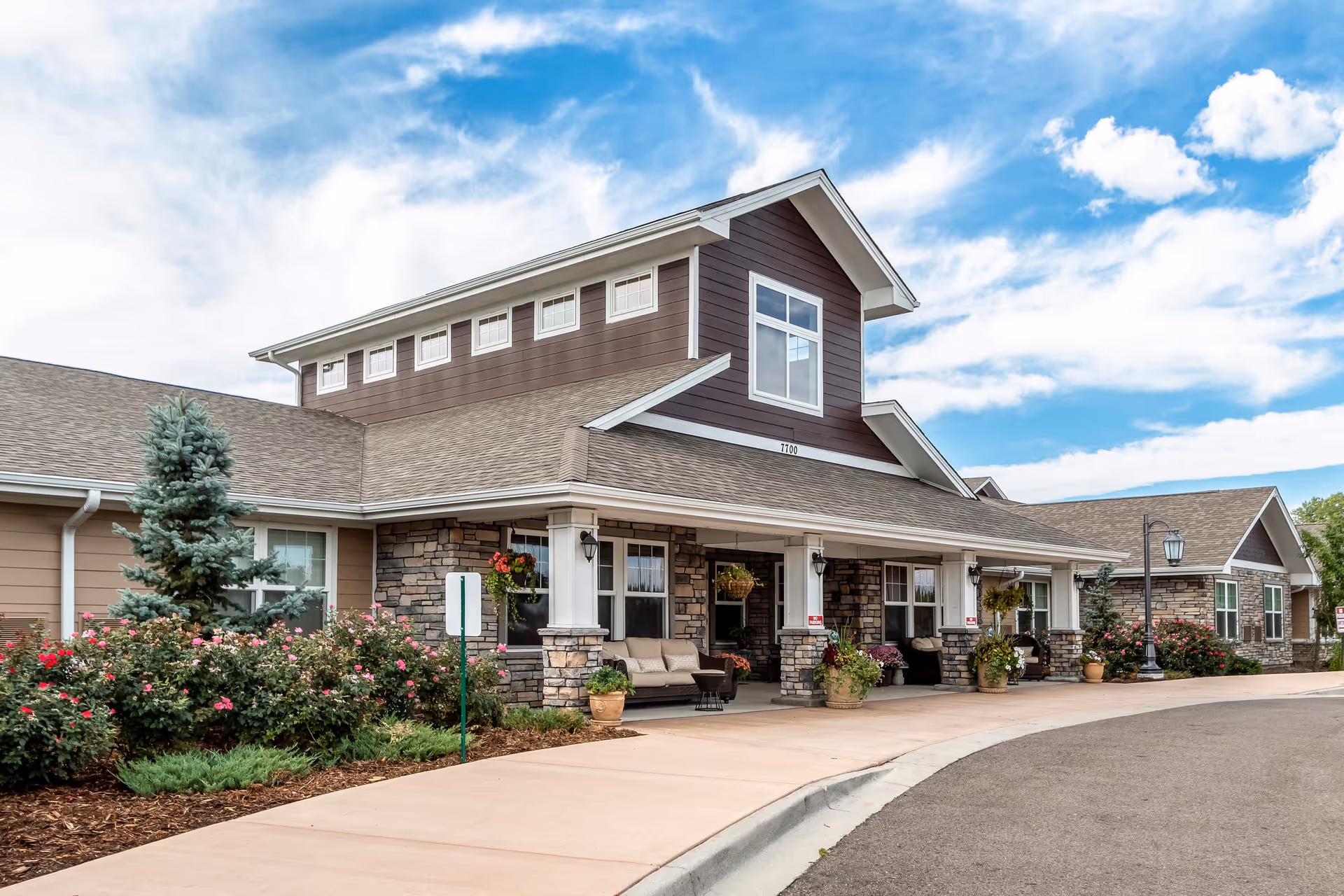 Exterior view of a senior living facility building with a covered porch, stone and wood siding, potted plants, and a curved driveway under a partly cloudy sky.