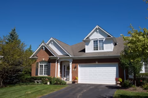 A brick and white-siding house with an attached two-car garage, driveway, and manicured front lawn under a clear blue sky.