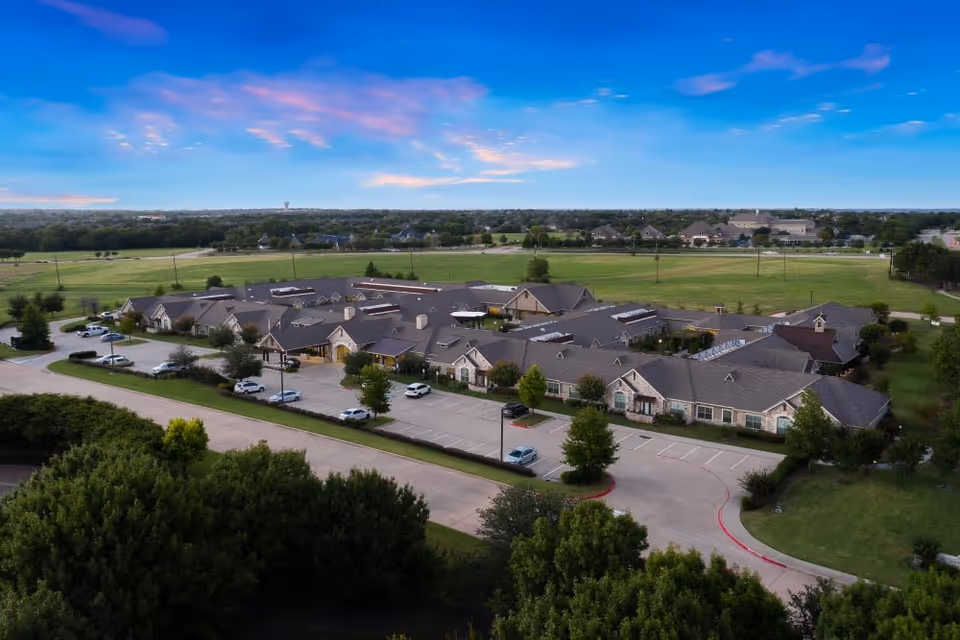 Aerial exterior view of a single-story senior living complex with parking lots, trees, and open green fields under a blue sky.