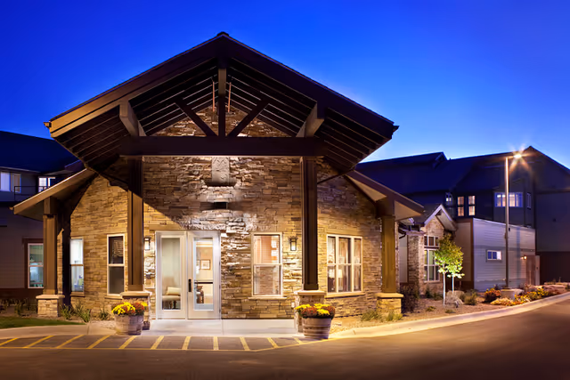 Illuminated stone-clad building entrance with a covered porte-cochere, potted flowers, and a curved driveway at dusk.