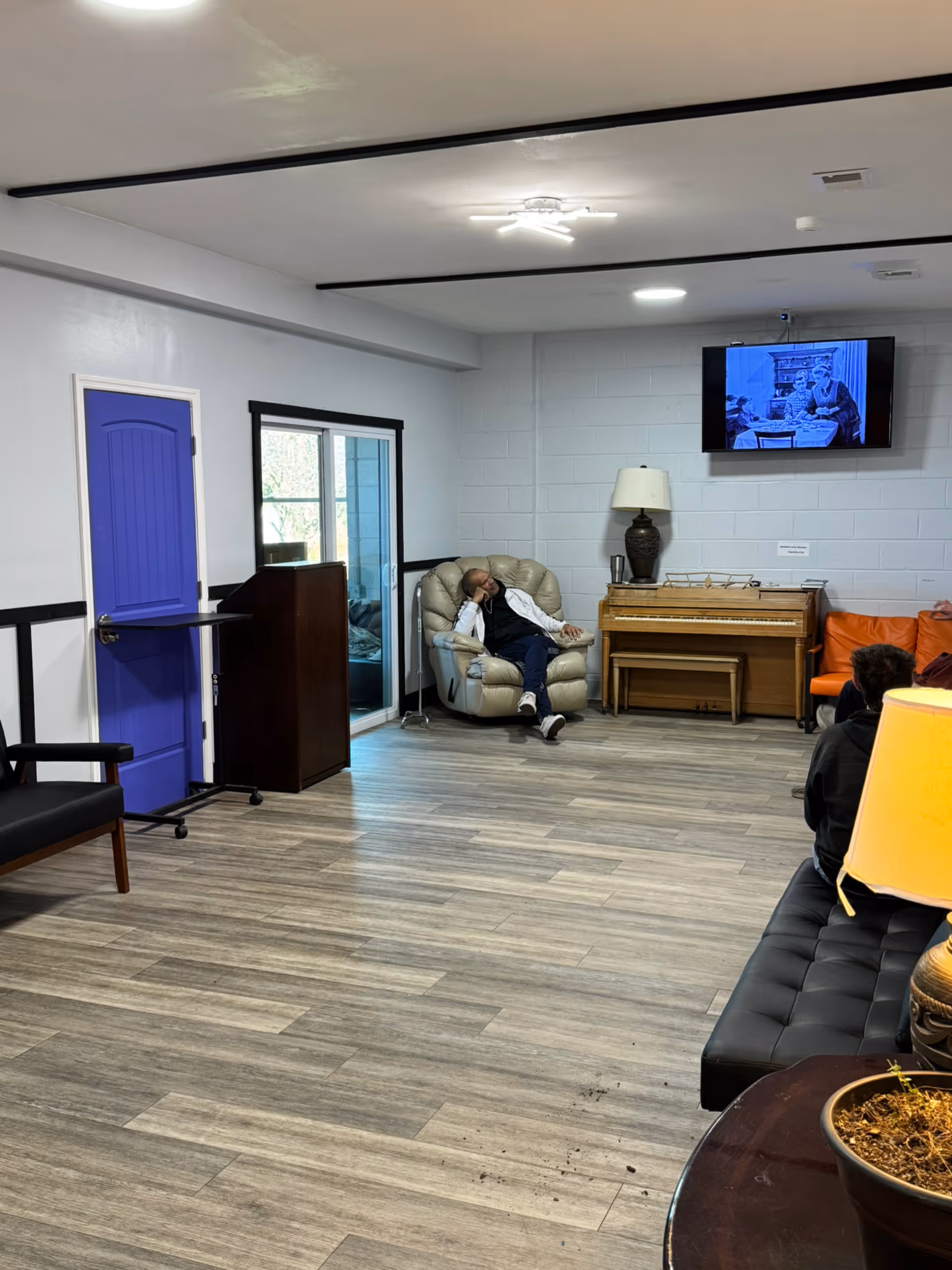 Residents' lounge with purple door, piano, couch, and a man resting in a recliner.