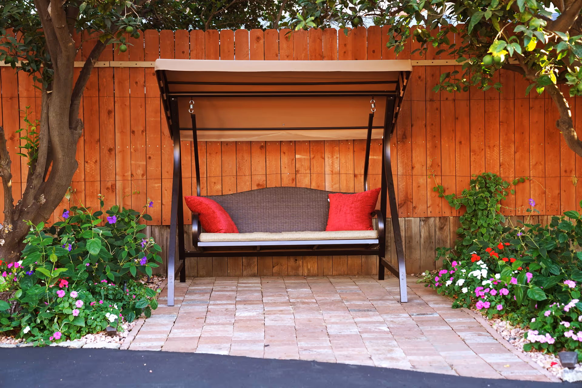 A brown outdoor swing with a canopy and two red cushions is placed on a paved area. The swing is set against a wooden fence, surrounded by green plants and colorful flowers on both sides. Two trees frame the scene.