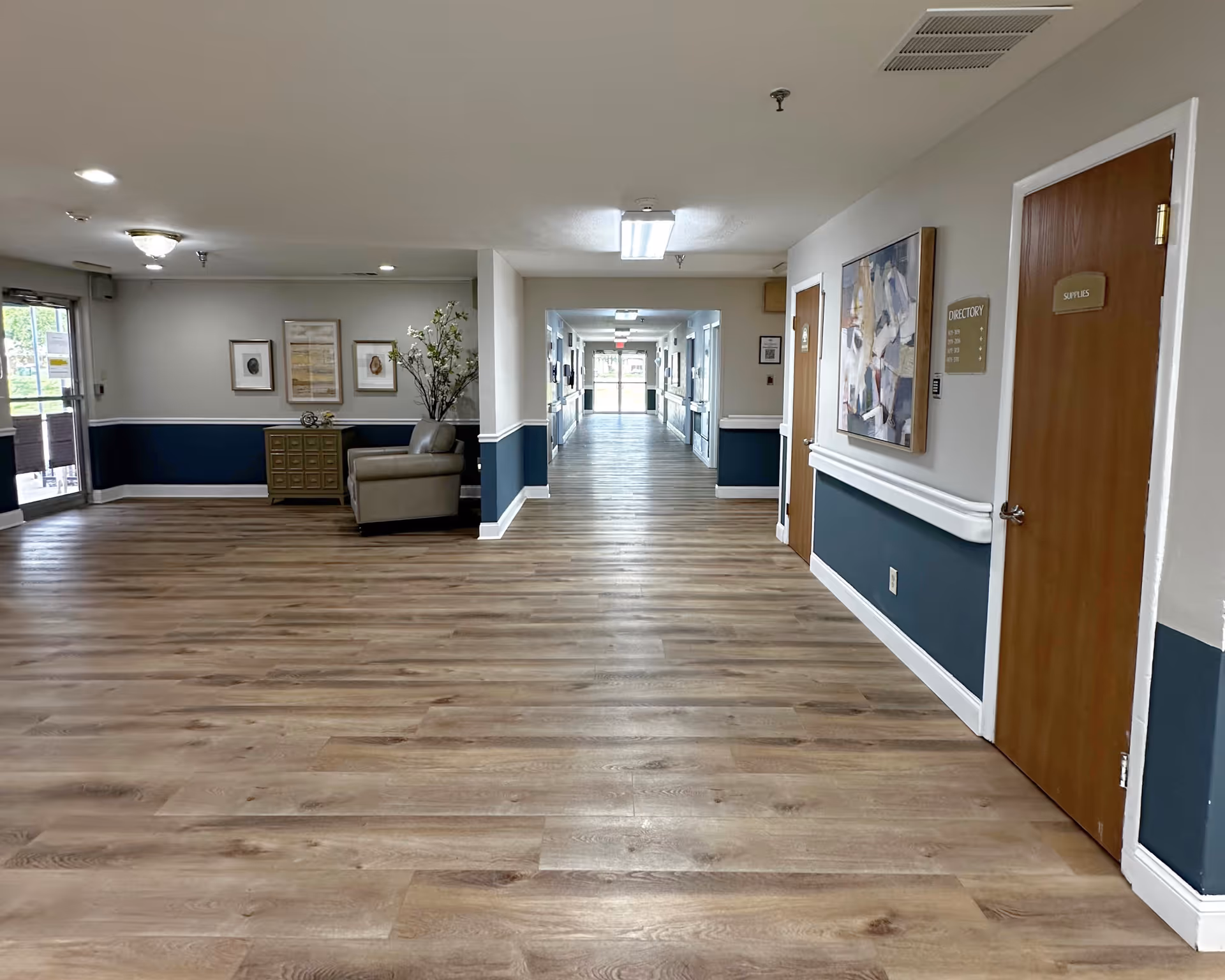 Wide interior corridor and lobby with wood-look flooring, seating, artwork, and doors lining the right wall.