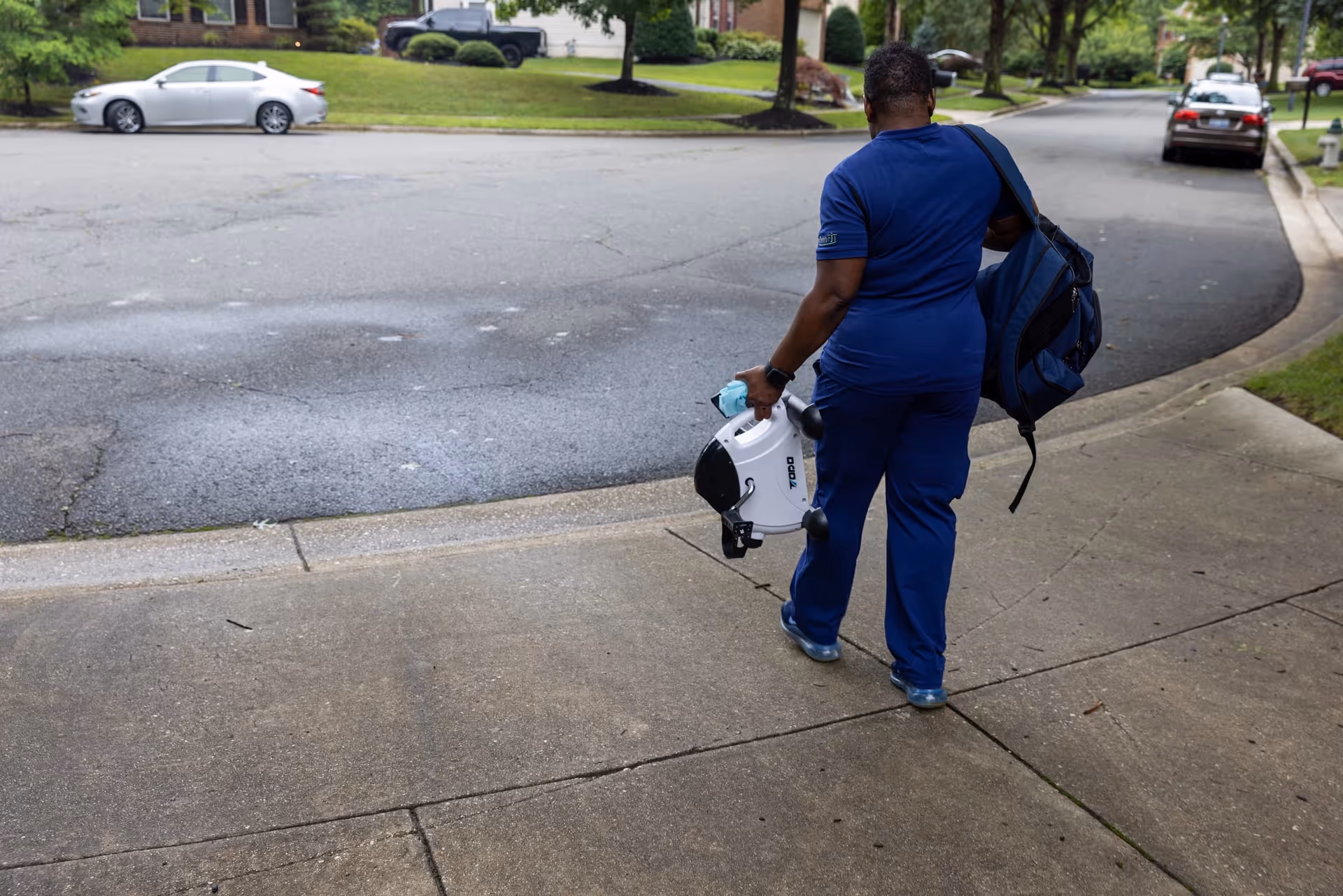 A caregiver in blue scrubs walks along a suburban street, carrying a bag and portable device.