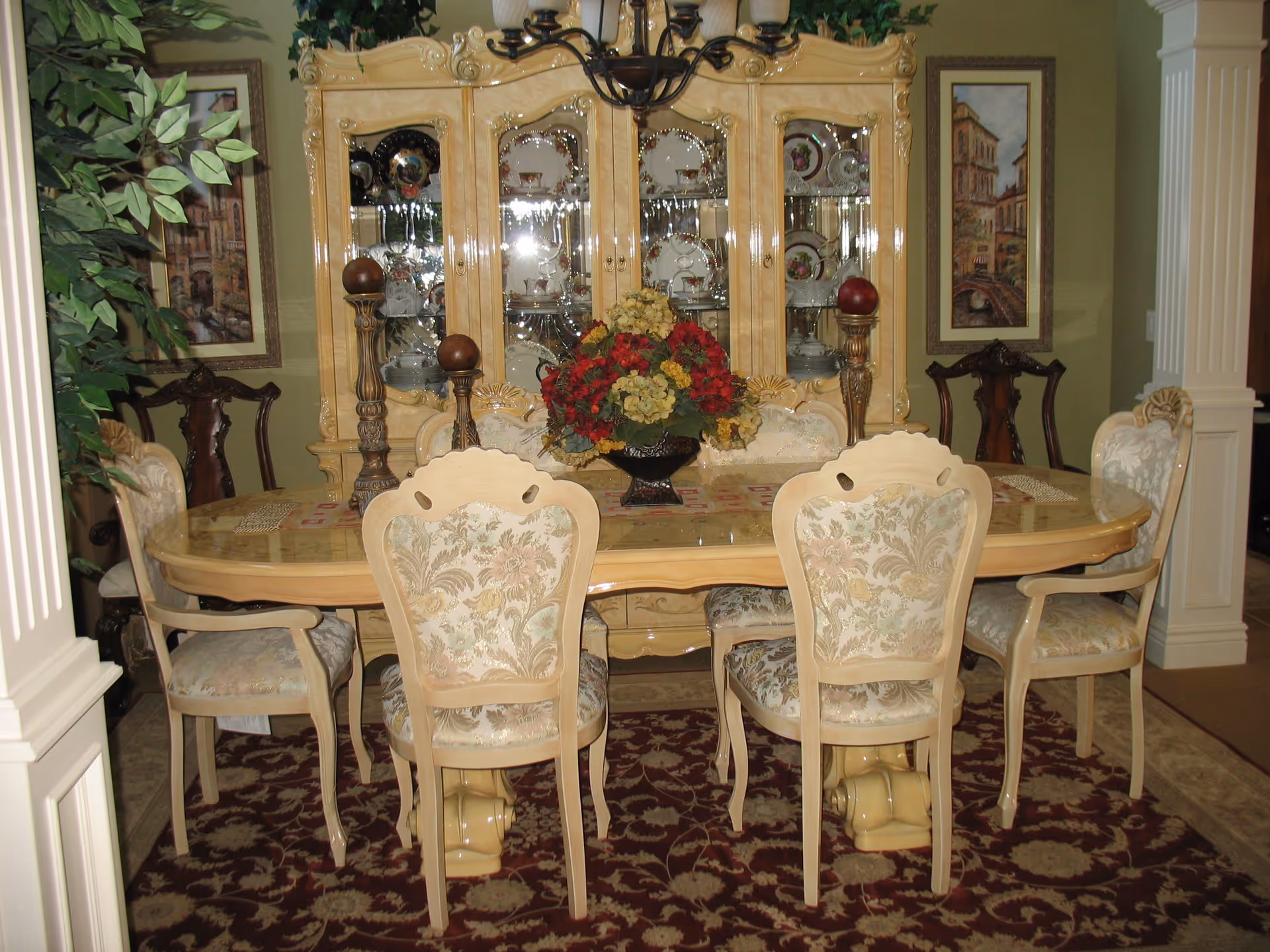 A formal dining room with a large wooden dining table surrounded by six upholstered chairs with floral patterns. The table has a centerpiece of red and yellow flowers and two tall decorative candle holders. Behind the table is a large wooden china cabinet displaying fine china plates and cups. The room is decorated with framed artwork on the walls and a large leafy plant on the left side. The floor has a patterned area rug under the table.