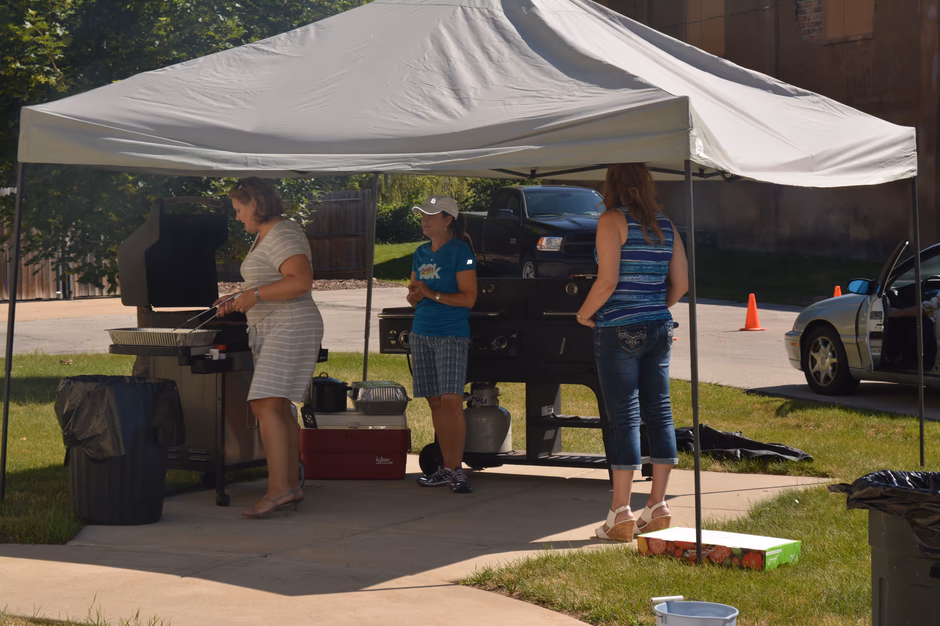 Three women grill under a white canopy in a grassy outdoor area.