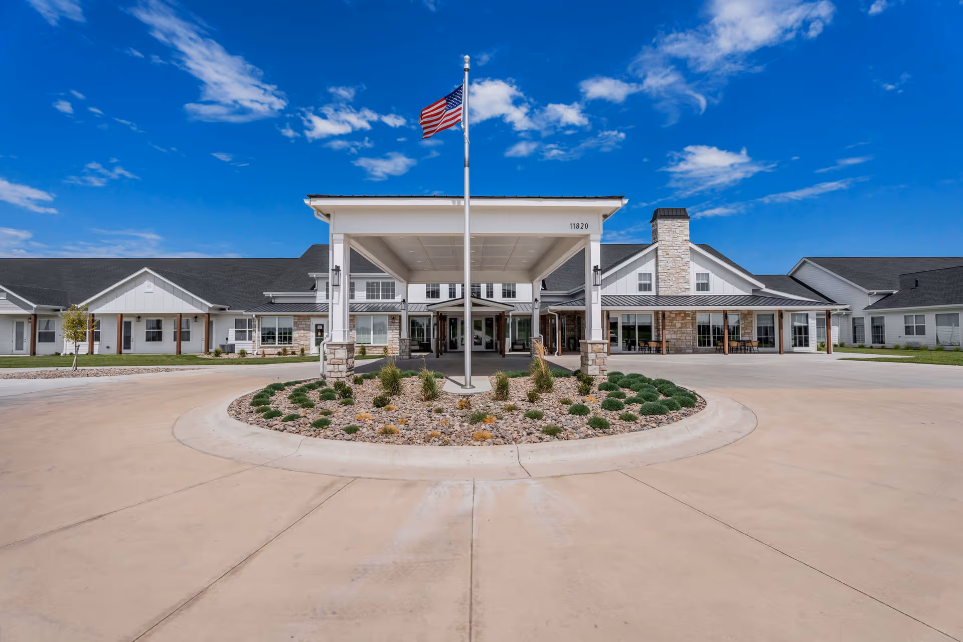 Front entrance of a single-story senior living facility with a covered porte-cochère, flagpole, circular driveway and landscaped island under a blue sky.