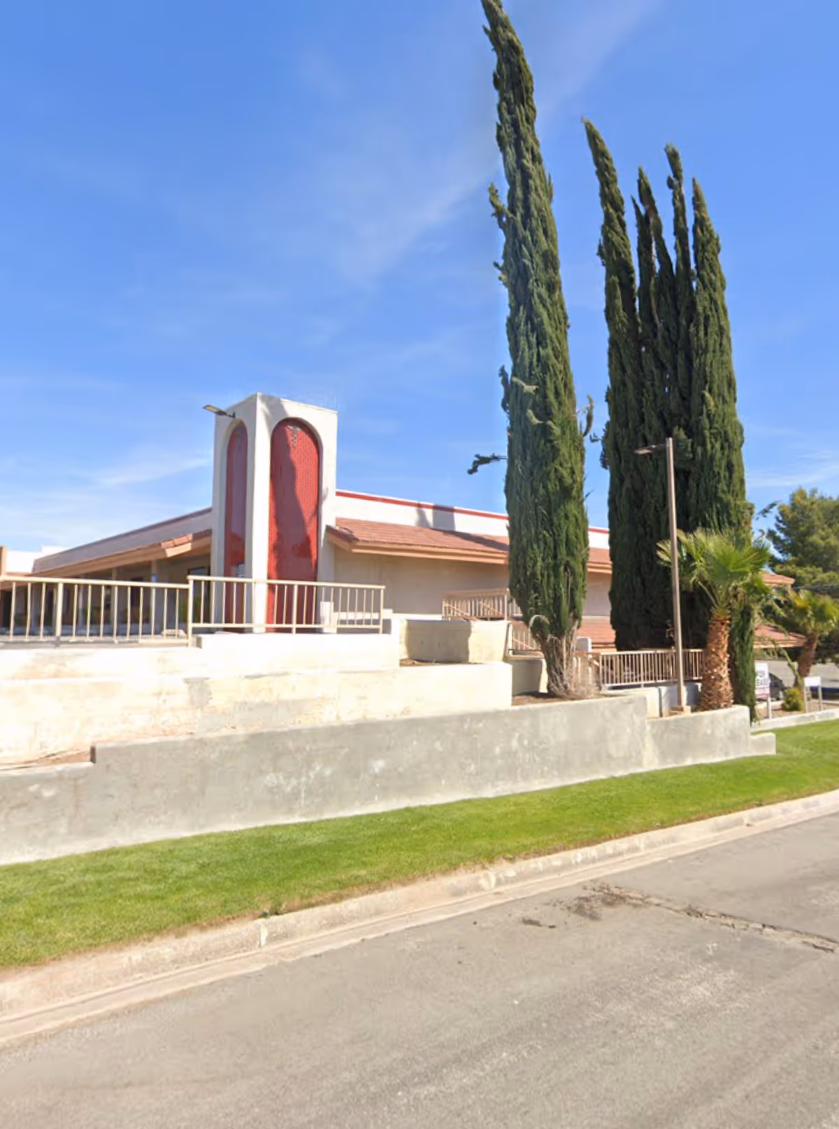 Exterior view of a facility with tall trees, a red entry arch, concrete steps, and lawn.