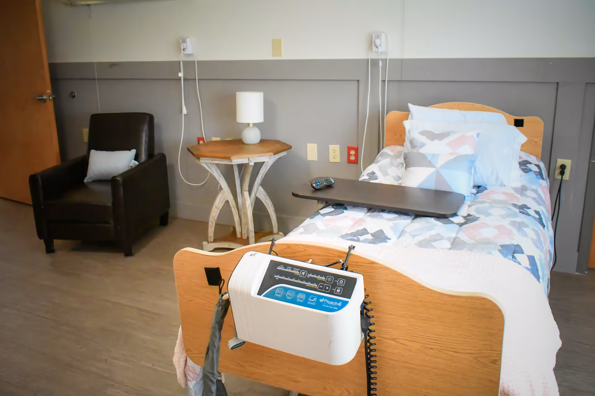 A senior living facility bedroom with a single bed featuring a colorful geometric patterned comforter and multiple pillows. A remote control rests on an overbed table. There is a medical device attached to the footboard of the bed. Next to the bed is a small wooden side table with a lamp, and a dark brown armchair with a small pillow is positioned nearby. The room has light-colored walls with a gray wainscoting and a wooden door partially visible on the left.