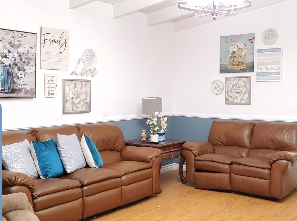 A cozy living room with two brown leather sofas adorned with blue and silver decorative pillows. The walls are painted white with a blue wainscoting and decorated with floral artwork and inspirational signs. A wooden side table with a lamp and a flower vase is placed between the sofas. The floor is wooden, and a modern chandelier hangs from the ceiling.